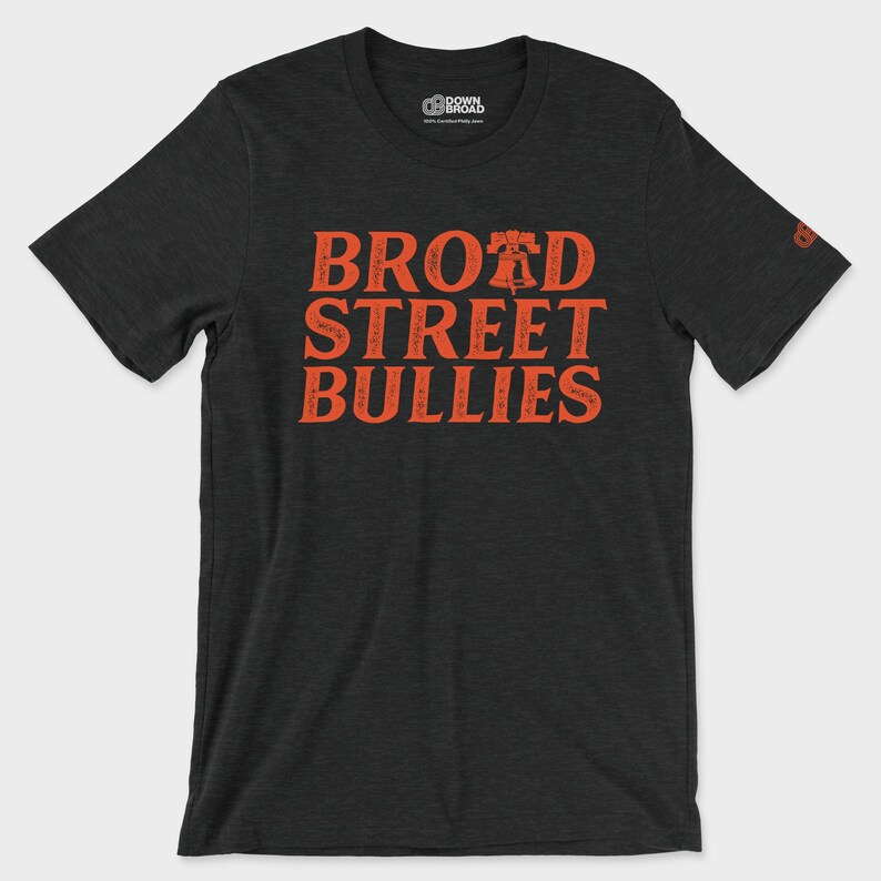 K&ouml;nnte beinhalten: Ein anthrazitfarbenes Kurzarm-T-Shirt mit dem orangefarbenen Schriftzug "BROAD STREET BULLIES", wobei ein Liberty Bell-Symbol den Buchstaben A in Broad ersetzt. Das T-Shirt hat einen Rundhalsausschnitt und ein kleines Logo am Innenkragen und am linken &Auml;rmel.