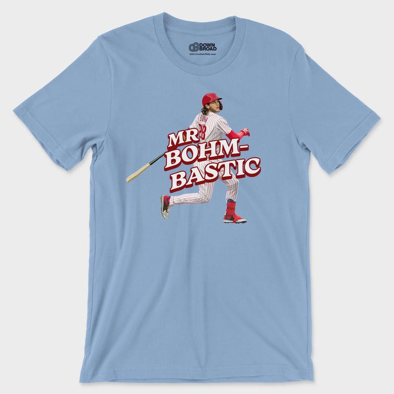 May include: A light blue short-sleeve t-shirt featuring a graphic of a baseball player in a red and white pinstripe uniform swinging a bat. The text "MR BOHM-BASTIC" is printed in bold red letters across the center of the graphic design.