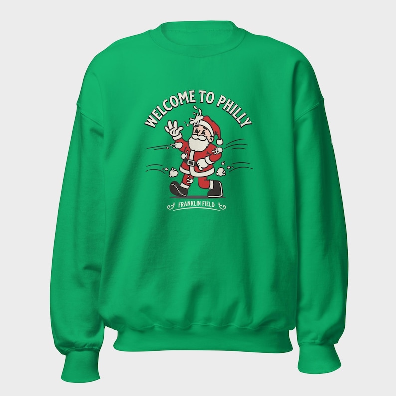 May include: A bright green crewneck sweatshirt featuring a graphic of Santa Claus waving, with the text "WELCOME TO PHILLY" arched above and "FRANKLIN FIELD" below. The design shows Santa being hit by snowballs in a cartoon style.