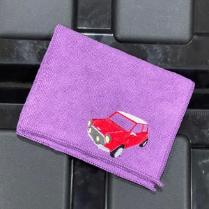 May include: A folded, rectangular, purple microfiber cloth featuring an embroidered red car design. The car has a white roof and black tyres. The cloth is likely for cleaning or detailing.