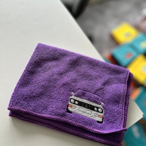 May include: A folded, purple microfiber cloth featuring an embroidered image of a white and silver car. The car has black details and orange lights. The cloth is on a white surface.