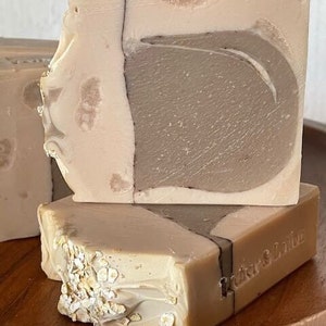 May include: Two bars of handmade soap, one with a brown and white swirl design and the other with oatmeal flakes. The bottom bar has the text "Cedar & Olive" printed on it.