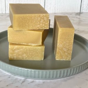 May include: Four bars of handmade soap stacked on a light grey ceramic plate. The soap is a light beige colour with a marbled pattern.