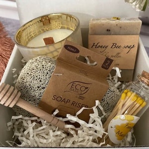 May include: A gift box with a candle in a gold glass, a bar of "Honey Bee soap", a wooden honey dipper, a loofah, a "Soap Riser" package, and a small glass jar of matches. The box is filled with white paper shreds.