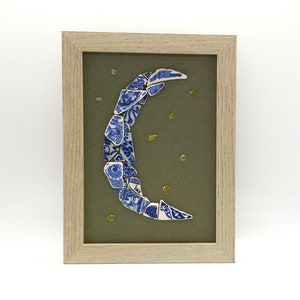 May include: A framed artwork featuring a crescent moon made of blue and white porcelain shards on a green background with gold glitter accents.