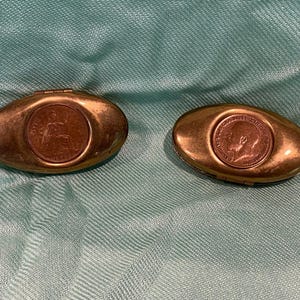 May include: Two antique brass oval pill boxes. Each box features a coin inset into the lid. The boxes are a warm brass colour and are set against a teal fabric background. The coins have detailed relief images.