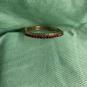 Pode incluir: Uma pulseira rígida articulada em tom dourado com uma fila contínua de pequenas pedras preciosas redondas de cor vermelho-escura na parte frontal. A banda metálica apresenta um intrincado padrão de filigrana vazada nas laterais e um fecho.
