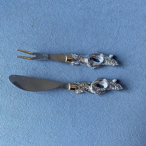 May include: A silver cheese fork and knife set. The handles feature an ornate fish design with gold accents. The fork has two tines, and the knife has a curved blade. The set is displayed on a blue surface.