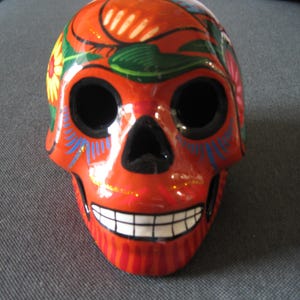 May include: A ceramic skull painted in bright orange with black eyes, a white smile, and colorful floral designs.