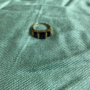 May include: A gold ring featuring three rectangular, deep red gemstones set in a row. The center stone is larger than the two flanking stones, and the band is a simple, polished gold metal. The ring is positioned on a textured, light green fabric.