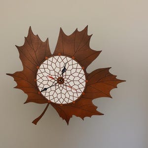 May include: A wooden wall clock shaped like a maple leaf. The clock face is a white geometric design with black and red hands. The leaf is brown with detailed veining.