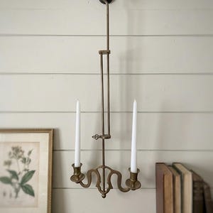 Puede incluir: Un candelabro de pared de latón con una varilla vertical ajustable, un gancho en la parte superior y dos brazos que se extienden hacia afuera para sostener dos velas cónicas blancas. El accesorio de metal tiene un acabado metálico y un mecanismo.
