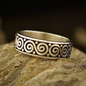 Puede incluir: Un anillo de banda de plata con un patrón repetitivo de espirales estilizados.