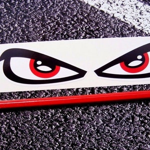 Puede incluir: Una hoja de dos pegatinas de vinilo blanco con contornos negros y ojos rojos. Los ojos tienen forma de ojo entrecerrado con una ceja negra y un iris rojo.