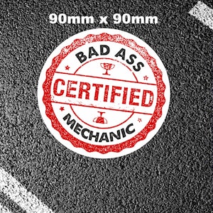 Könnte beinhalten: Ein runder Aufkleber in Rot und Weiß mit dem Text "Bad Ass Certified Mechanic" in einer verwitterten Schriftart. Der Aufkleber ist 90 mm x 90 mm groß und befindet sich auf einem schwarzen Asphalt-Hintergrund.