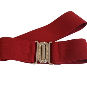 May include: A wide, red elastic belt with a silver-colored, rectangular buckle. The buckle has a decorative, vertical row of small, clear embellishments. The belt is designed to be worn around the waist.