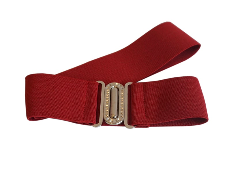 May include: A wide, red elastic belt with a silver-colored, rectangular buckle. The buckle has a decorative, vertical row of small, clear embellishments. The belt is designed to be worn around the waist.