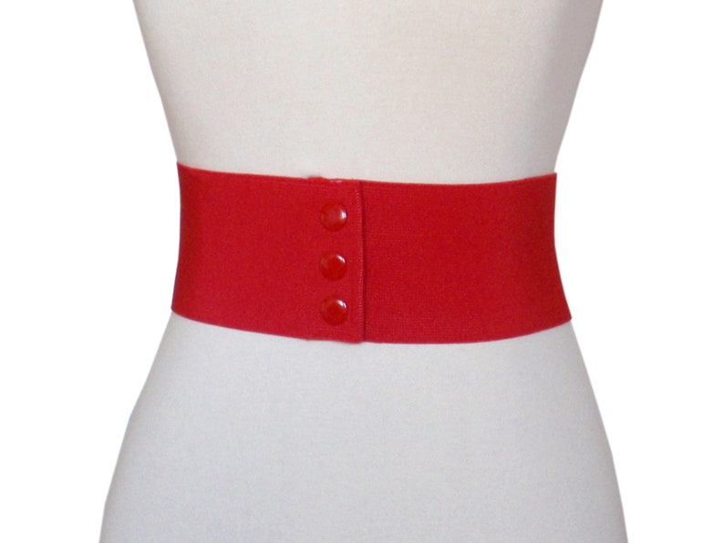 May include: A red elastic belt with three buttons. The belt is wide and has a smooth, matte finish.