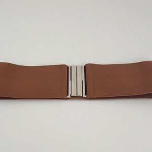 May include: A brown elastic belt with a silver buckle. The belt is flat and wide, and the buckle is simple and understated.