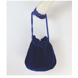 Op de afbeelding: Een marineblauwe fluwelen tas met trekkoord en een bijpassende touwriem. De tas wordt door een hand vastgehouden, wat de zachte textuur en het elegante ontwerp benadrukt. De opening van de tas is gerimpeld met bijpassende blauwe stof.