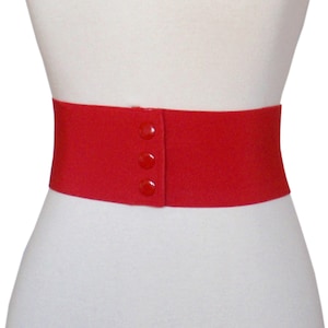 Op de afbeelding: Een rode elastische riem met drie knopen. De riem is breed en heeft een gladde, matte afwerking.