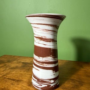 Könnte beinhalten: Eine zylindrische Vase mit einem marmorierten Muster aus weißen und rotbraunen Wirbeln. Das Gefäß hat eine glatte, glänzende Oberfläche mit einem leicht ausgestellten Rand an der Oberseite und einem geraden Körper, der sich zur Basis hin verjüngt.