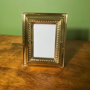 May include: A rectangular picture frame with a polished gold finish. The frame features a smooth outer border and an inner decorative band with a repeating textured pattern surrounding a blank white rectangular center insert for a photograph.