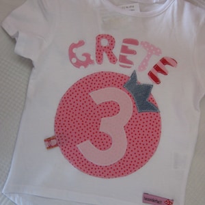Könnte beinhalten: Weißes T-Shirt mit einer rosa und roten Applikation des Wortes "GREY" und einer Zahl "3" in einem Kreis mit rosa Punkten. Das Shirt hat auch ein kleines Etikett mit der Aufschrift "besonderheit".