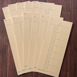 May include: A stack of beige time cards with green lines and text. The top card has the text "NAME" and "WEEK ENDING" at the top. The card is divided into columns for each day of the week, with the days of the week listed in green text. The text "IN" and "OUT" are listed in each column. The bottom card has the text "CTOPS 1200 Replaces K 14-101".