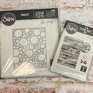 May include: Two Sizzix Tim Holtz crafting tools in clear plastic packaging. The left package is a Thinlits die with a circular bubble pattern, item 665211. The right package is a 3-D Texture Fades mini embossing folder with a wood grain pattern, item 665460.