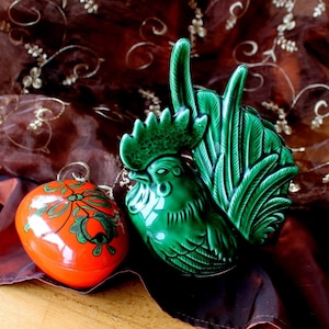 Op de afbeelding: Een groen keramiek haanbeeldje met een bruine en gouden stoffen achtergrond met patroon. Een rood en groen geverfd ei-vormig doosje is ook in de afbeelding.