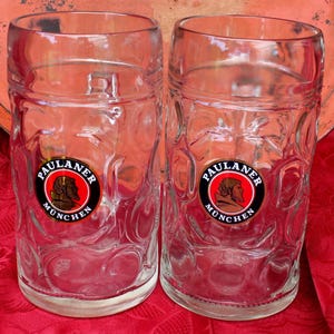 Könnte beinhalten: Zwei klare Bierkrüge mit dem Logo "Paulaner München" vor rotem Hintergrund. Die Krüge haben ein strukturiertes Design und sind leer. Im Hintergrund befindet sich ein runder, rostfarbener Tablett.