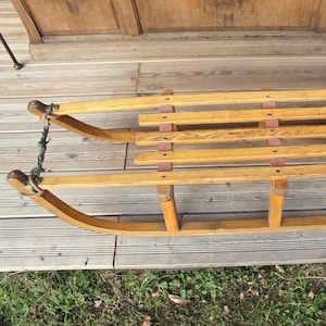 Op de afbeelding: Een houten slee met een touw aan de voorkant. De slee heeft een houten zitting met een label waarop "Made in Austria" staat.