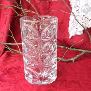 Könnte beinhalten: Eine klare, rechteckige Glasvase mit Blättermuster. Die Vase steht auf einer roten Tischdecke mit Blumenmuster. Zweige sind in der Nähe der Vase platziert. Eine weiße Spitzendecke befindet sich im Hintergrund.