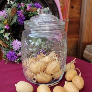 Può includere: Un barattolo di vetro con un coperchio, pieno di uova di legno. Il barattolo ha una superficie testurizzata e si trova su una tovaglia rossa. Le uova di legno sono sparse intorno al barattolo sulla tovaglia.