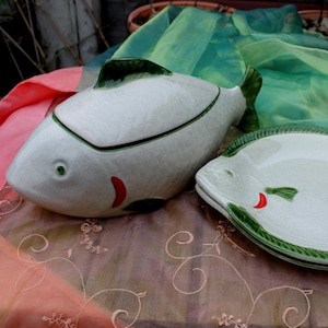 Op de afbeelding: Een keramische visvormige serveerschaal en een ovenschotel met deksel. Beide zijn wit met groene accenten, waaronder vinnen en een decoratieve rand. Rode accenten worden gebruikt voor de mond. De gerechten staan op een patroon.