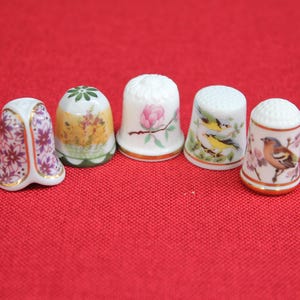 May include: Five ceramic thimbles arranged in a row. The first features a purple floral pattern with gold trim. The second has a yellow landscape design. The third displays a pink flower. The fourth shows two yellow birds. The fifth depicts a bird.