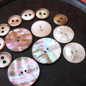Peut inclure: Une collection de 11 boutons en nacre de différentes tailles et couleurs. Les boutons ont deux trous et sont disposés en demi-cercle sur une surface noire.