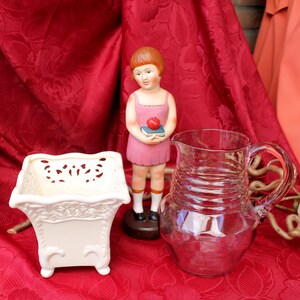 Peut inclure: Une nature morte composée d'un pot carré crème avec des découpes décoratives, d'une cruche en verre vintage et d'une figurine d'une fille en robe rose tenant un livre et une pomme. Les objets sont posés sur un fond de tissu rouge.