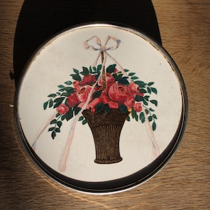 May include: A round, white ceramic trinket box with a silver rim. The lid features a painted design of a basket of pink roses with green leaves and a white ribbon.