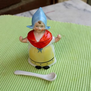 May include: A ceramic figurine of a woman in traditional Dutch attire, featuring a blue bonnet, red shawl, and yellow apron. A small white spoon rests nearby. The figurine has black shoes and is on a green surface.