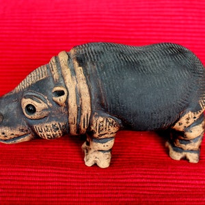 Könnte beinhalten: Eine kleine, schwarze Keramik-Nilpferd-Figur mit braunen Akzenten. Die Figur hat eine strukturierte Oberfläche und steht auf einem roten Hintergrund.