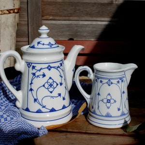 Peut inclure: Un ensemble de théière et de crémier blancs avec des motifs floraux bleus. La théière a un bec verseur et une poignée. Le crémier a une poignée et un bec verseur. Les deux pièces sont posées sur une surface en bois.