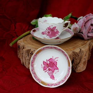 Può includere: Un set di tazze da tè e piattini in porcellana rosa e bianca con un motivo floreale. Le tazze e i piattini hanno un bordo delicato e ondulato. Il motivo floreale è di colore rosa e presenta un fiore stilizzato con foglie. Il set è esposto su una superficie in legno.