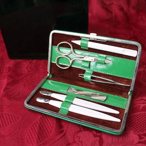 Könnte beinhalten: Ein grünes Reiseetui aus Kunstleder mit braunem Innenfutter, das ein Set Maniküre-Werkzeuge enthält. Die Werkzeuge umfassen eine Nagelschere, eine Nagelfeile, einen Nagelhaut-Schieber und eine Pinzette.