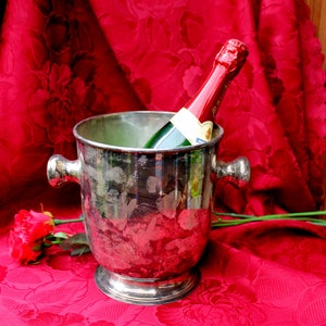 Puede incluir: Un cubo de champán plateado con una botella de champán dentro. El cubo está sobre un mantel rojo con una rosa roja.