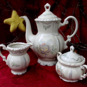 Op de afbeelding: Een wit porseleinen theeservies met bloemenmotieven. De set bevat een theepot, een roomkan en een suikerpot.