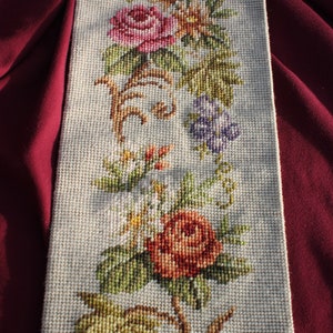 Op de afbeelding: Een beige stoffen paneel met een borduurwerk met bloemenmotief. De borduurwerk bevat roze, rode, oranje, gele, groene en paarse bloemen en bladeren.