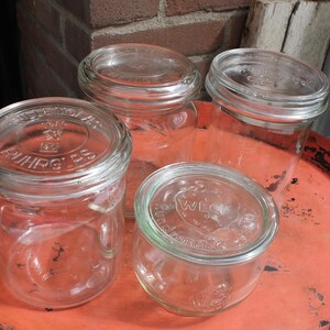 Op de afbeelding: Vier heldere glazen potten met deksels, van verschillende maten, zijn gerangschikt op een verweerde oranje metalen dienblad. De potten hebben reliëftekst op de deksels. De lade heeft een versleten, rustieke uitstraling.