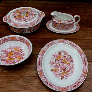 Può includere: Un set di stoviglie in ceramica con base bianca e un motivo floreale rosso e giallo. Il set comprende una zuppiera con coperchio, una salsiera su un piattino, una ciotola rotonda e un piatto da portata ovale, tutti con un bordo decorativo rosso.
